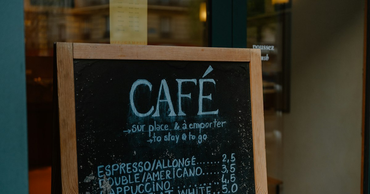 A hand-chalked café sign on a wooden easel outside a shop, listing coffee prices.