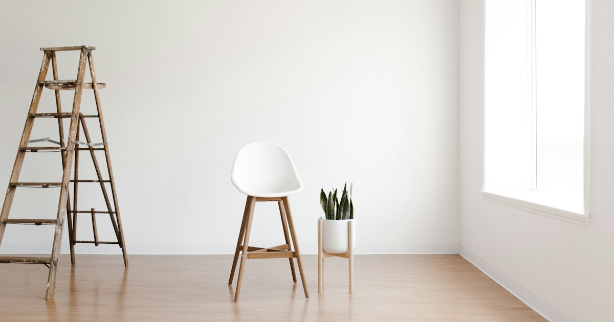 An empty white stool in a bare, bright photo studio, beside a potted plant and a wooden stepladder.