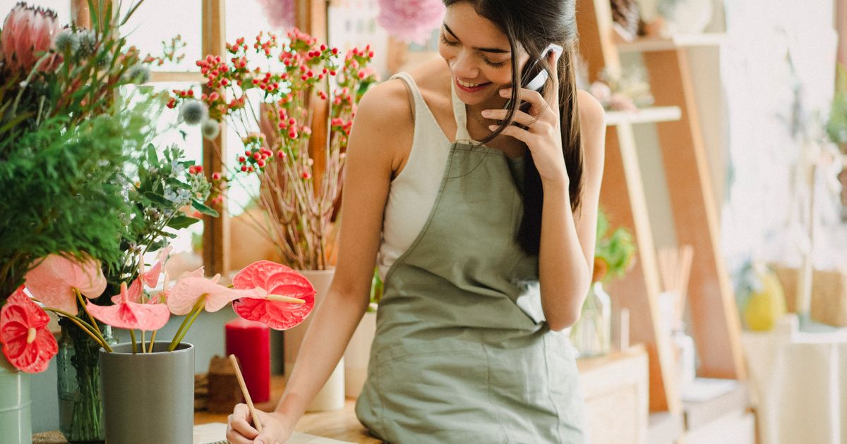 A florist in an apron smiling as she takes a phone call, writing a note at her counter among potted flowers.