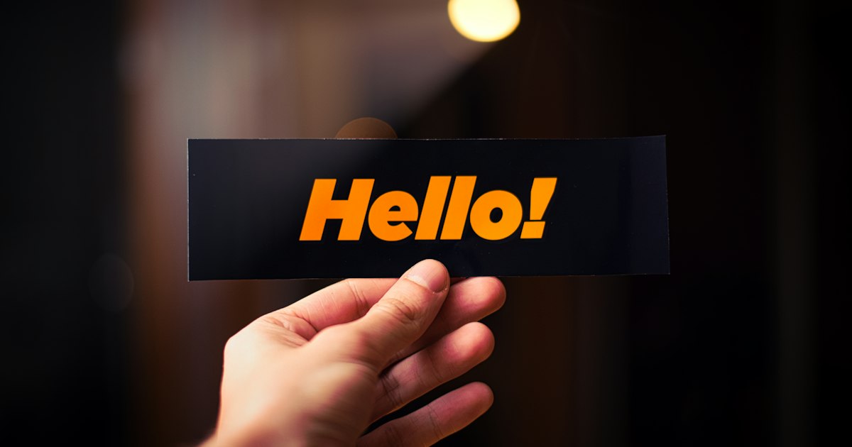 A hand holding up a black sticker that reads "Hello!" in bright orange letters.