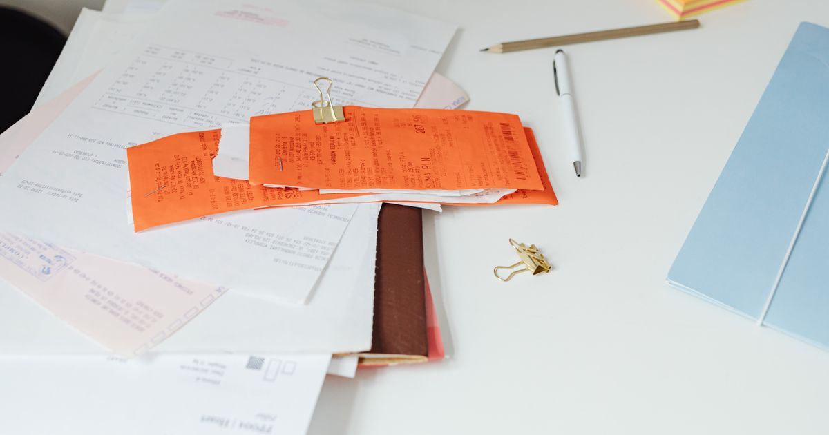 A pile of printed invoices and receipts held with a gold clip on a white desk, beside a pen and pencil.
