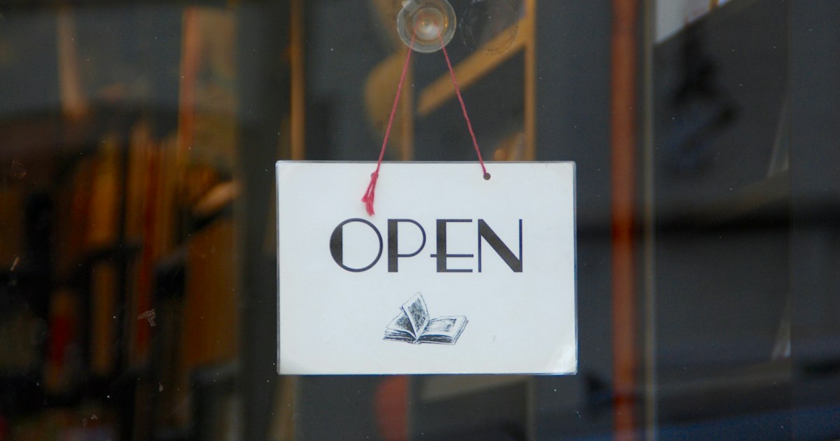 A white OPEN sign hanging on red string inside the glass door of a bookshop.