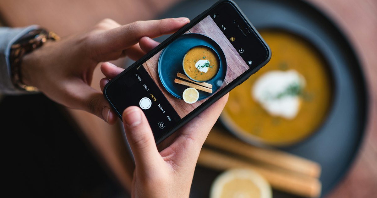 Hands holding a phone above a bowl of soup, taking a photo of it from directly overhead.