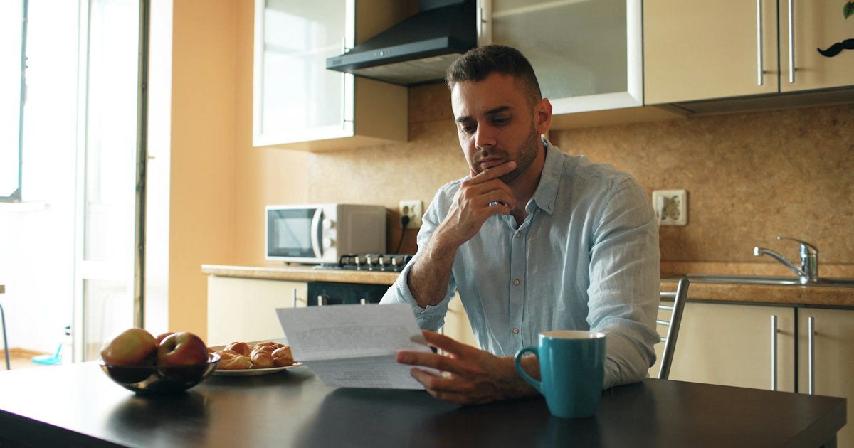 A man at a kitchen table reading a letter with a frown, a mug of tea beside him.