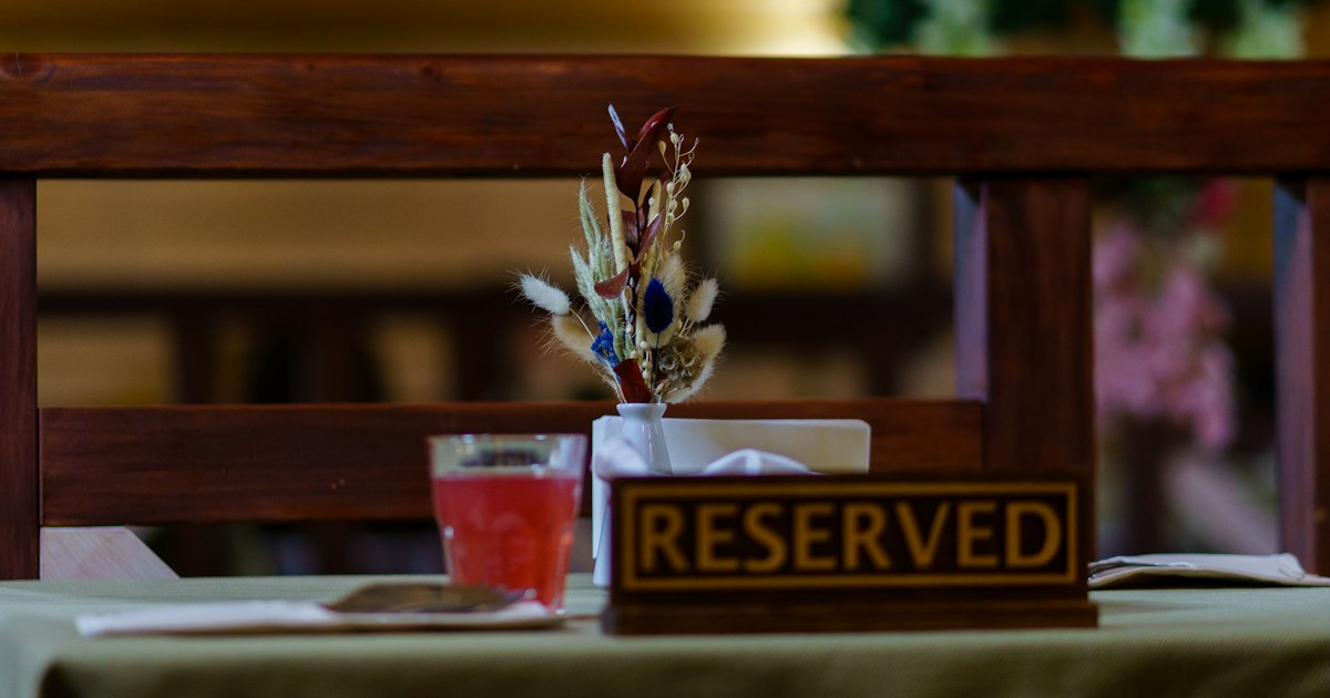 A small wooden RESERVED sign on a restaurant table beside a glass and a little vase of dried flowers.